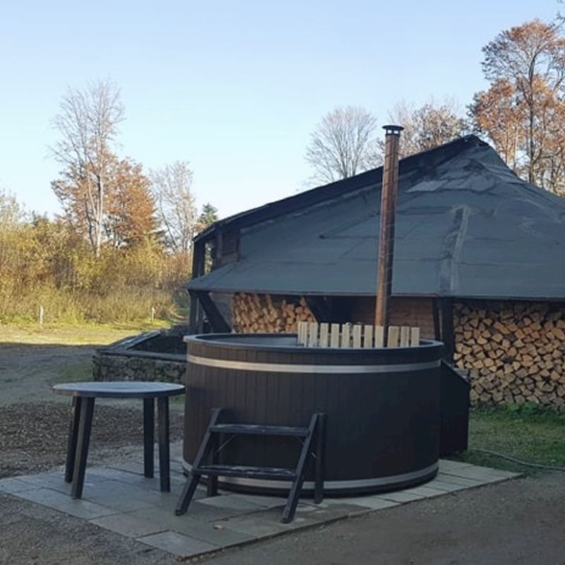 Drevený kúpací sud s komínom pod prístreškom, vedľa naukladané drevo a jesenné stromy.
